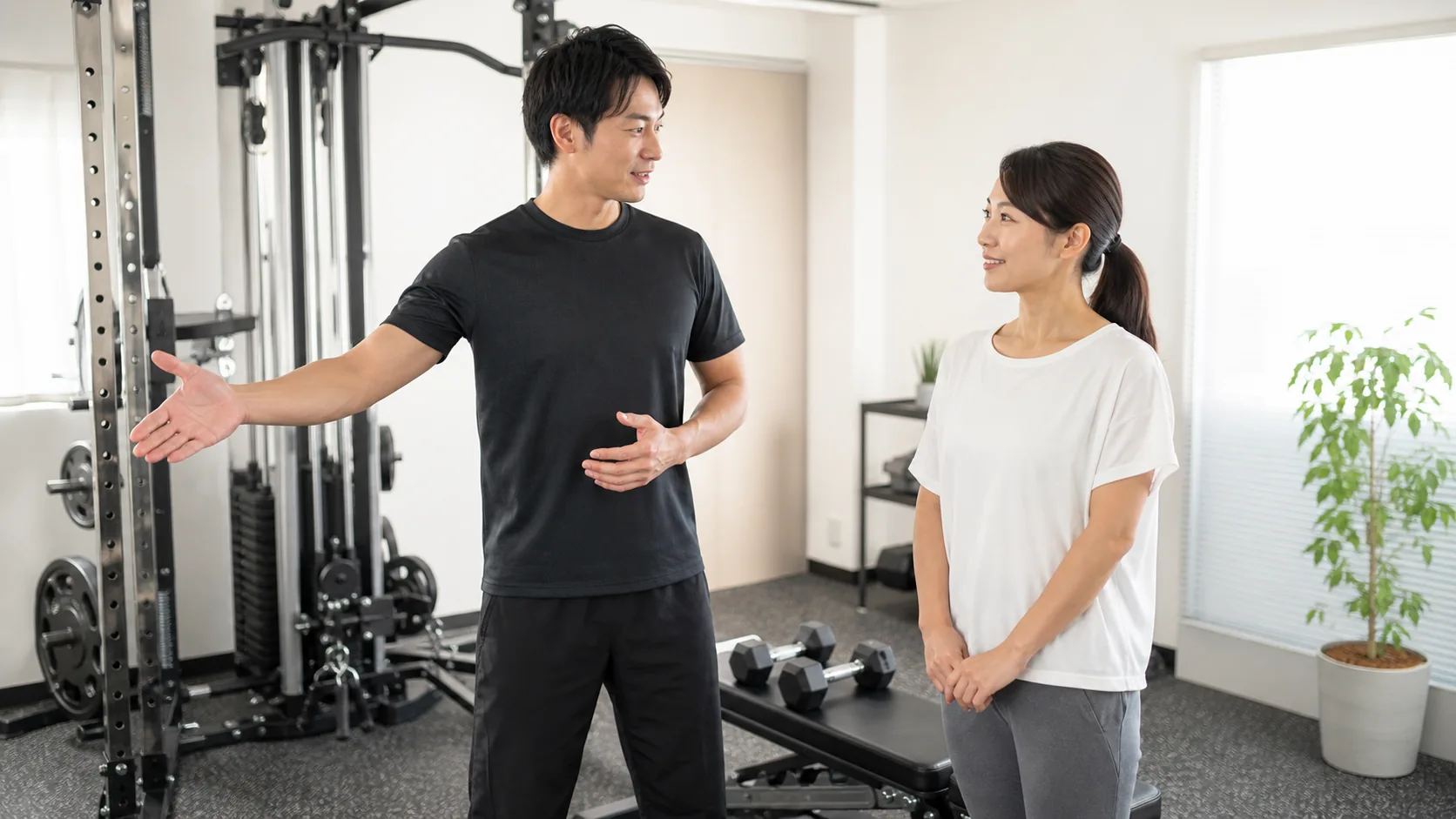 清潔なパーソナルジムで初心者の日本人女性がトレーナーから丁寧な説明を受ける様子